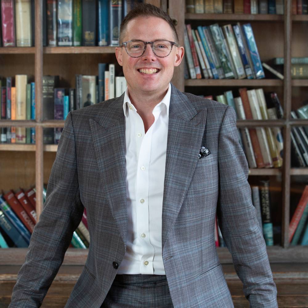 Ben Plummer-Powell smiling at the camera with a bookshelf behind him.