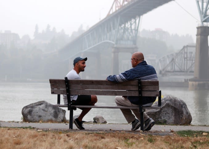 Alex speaks with a man being interviewed for Emergence Out of the Shadows on a bench by a river